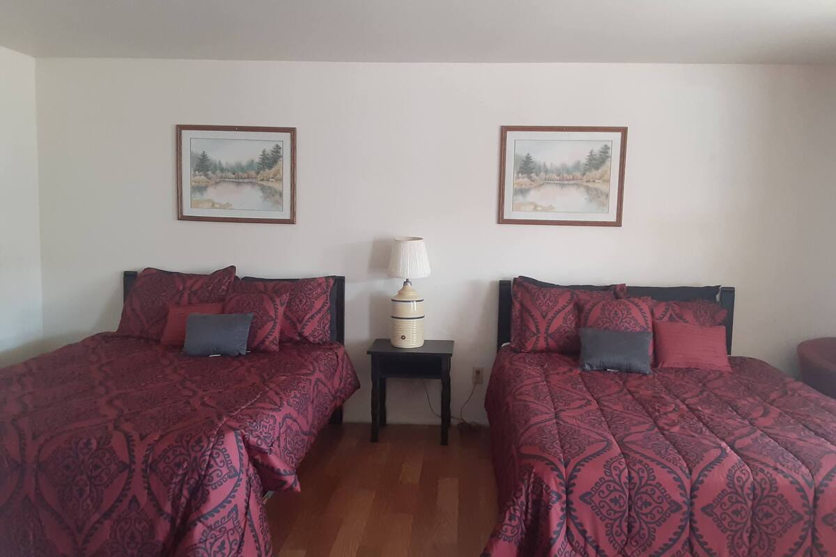 A room with two beds, each with a patterned bedspread and pillows, a bedside table with a lamp, and two framed pictures on the wall.