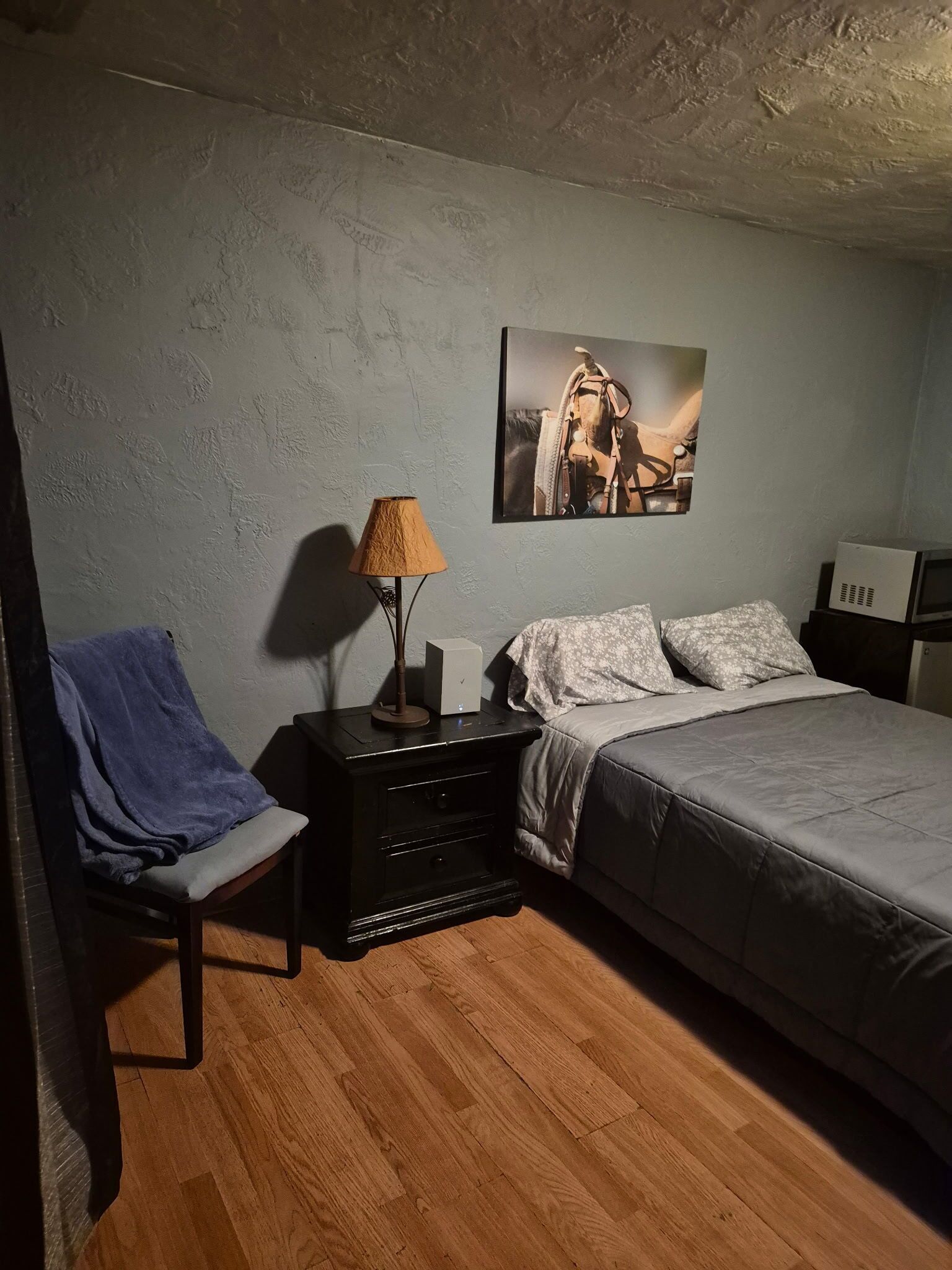 A bedroom with a bed, a nightstand, a lamp, a chair with a blanket, a picture on the wall, and a microwave.
