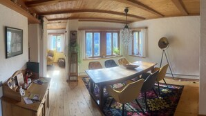 Tilava huone, jossa on puinen ruokapöytä, tuoleja ja kattokruunu. Huoneessa on ikkunat, joissa on verhot, puinen katto ja pieni sivupöytä, jonka päällä on maljakko.