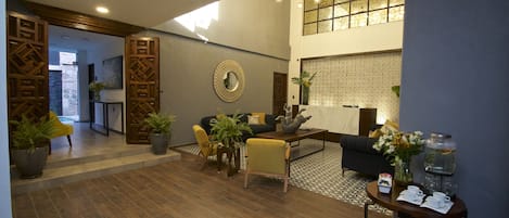 A spacious living area with a wooden floor, a sofa, a coffee table, and a dining table with chairs. There is a decorative mirror on the wall and a potted plant on the floor.
