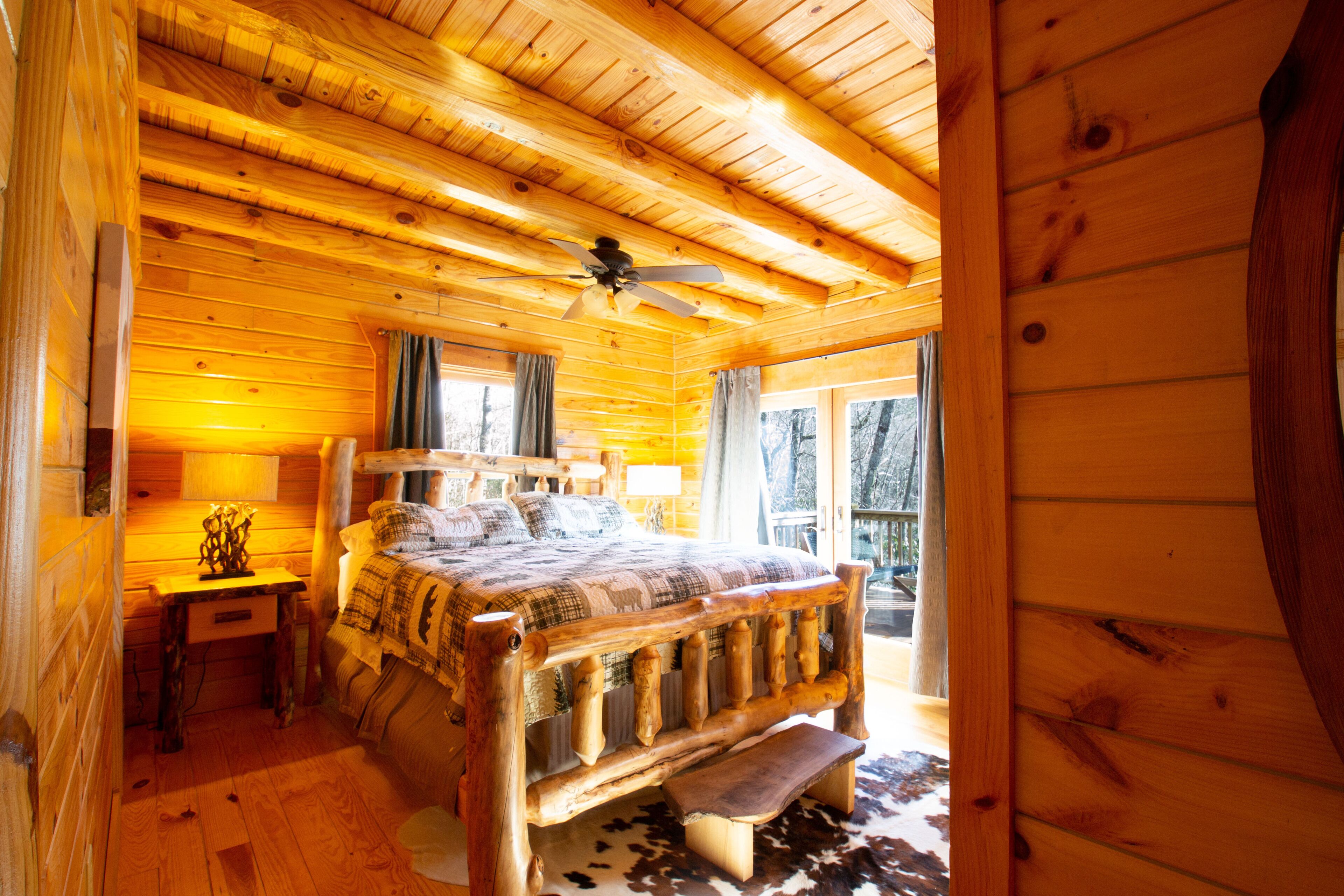Une chambre dans une cabane en bois, avec un lit, une table de chevet, un ventilateur de plafond et une fenêtre avec des rideaux.