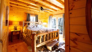 Un dormitorio de cabaña de madera con una cama, una mesita de noche, un ventilador de techo y una ventana con cortinas.