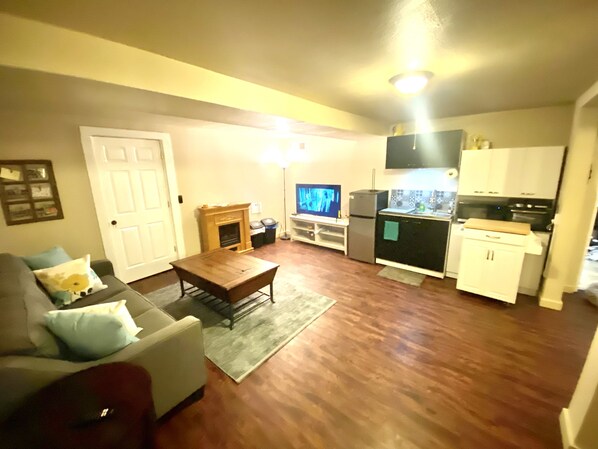 Un espacio habitable compacto con sofá, mesa de centro, chimenea, televisor, zona de cocina con armarios y electrodomésticos, y una puerta.