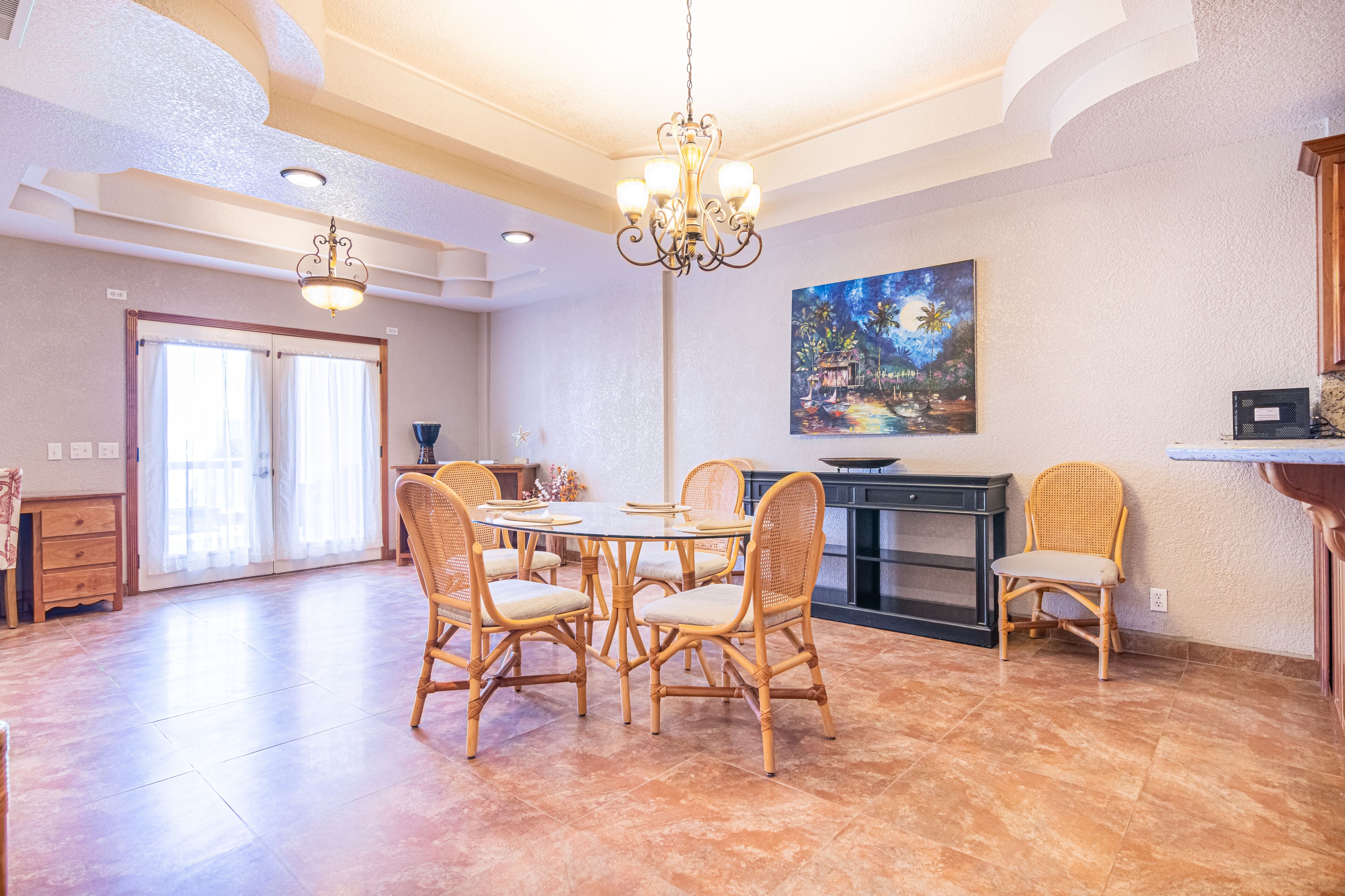 Une salle à manger spacieuse avec une table ronde et six chaises en osier, un lustre et un tableau accroché au mur.