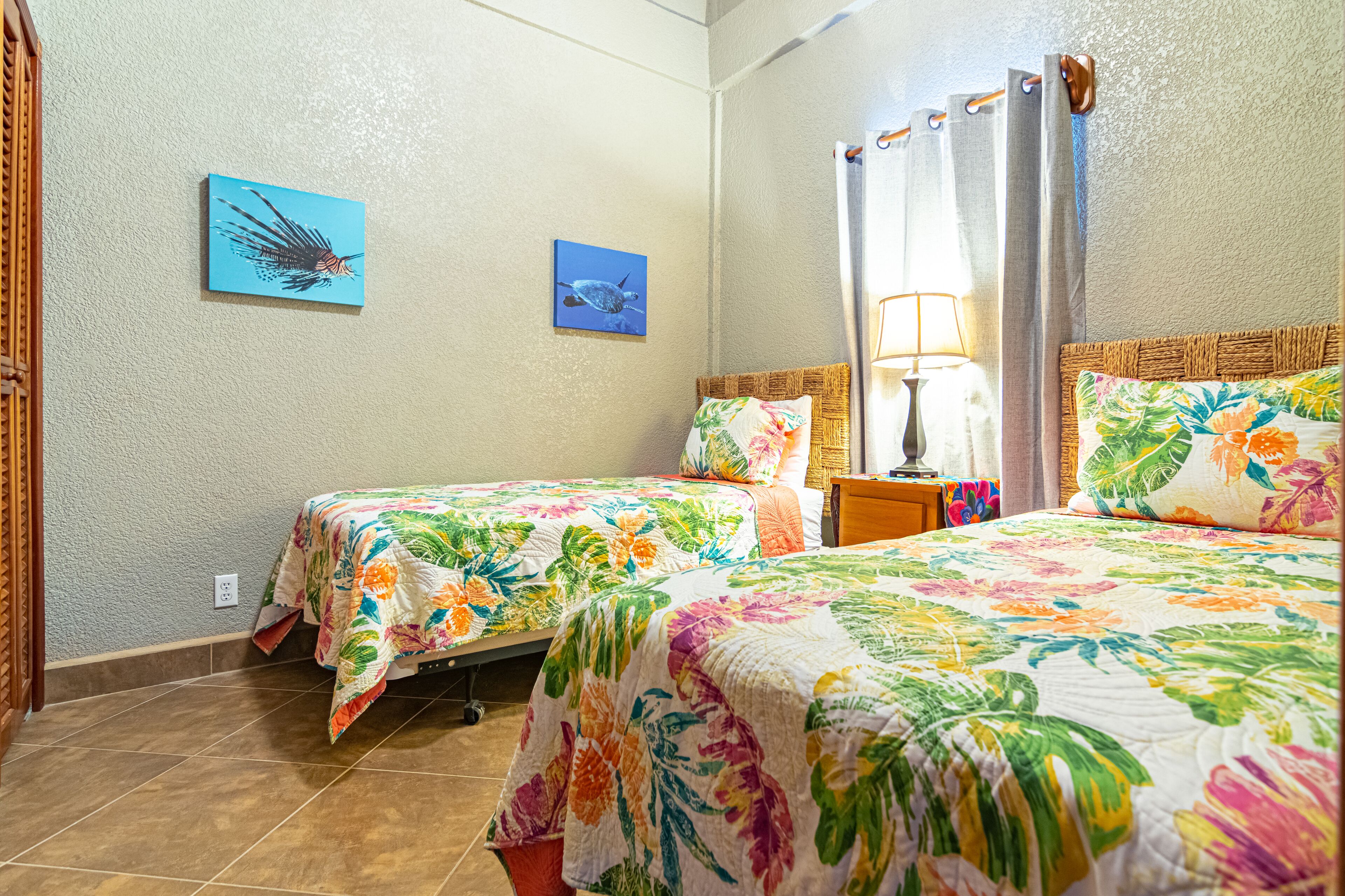 Une chambre d’hôtel avec deux lits, des couvre-lits à motifs floraux, une table de chevet avec une lampe et deux œuvres d’art murales.