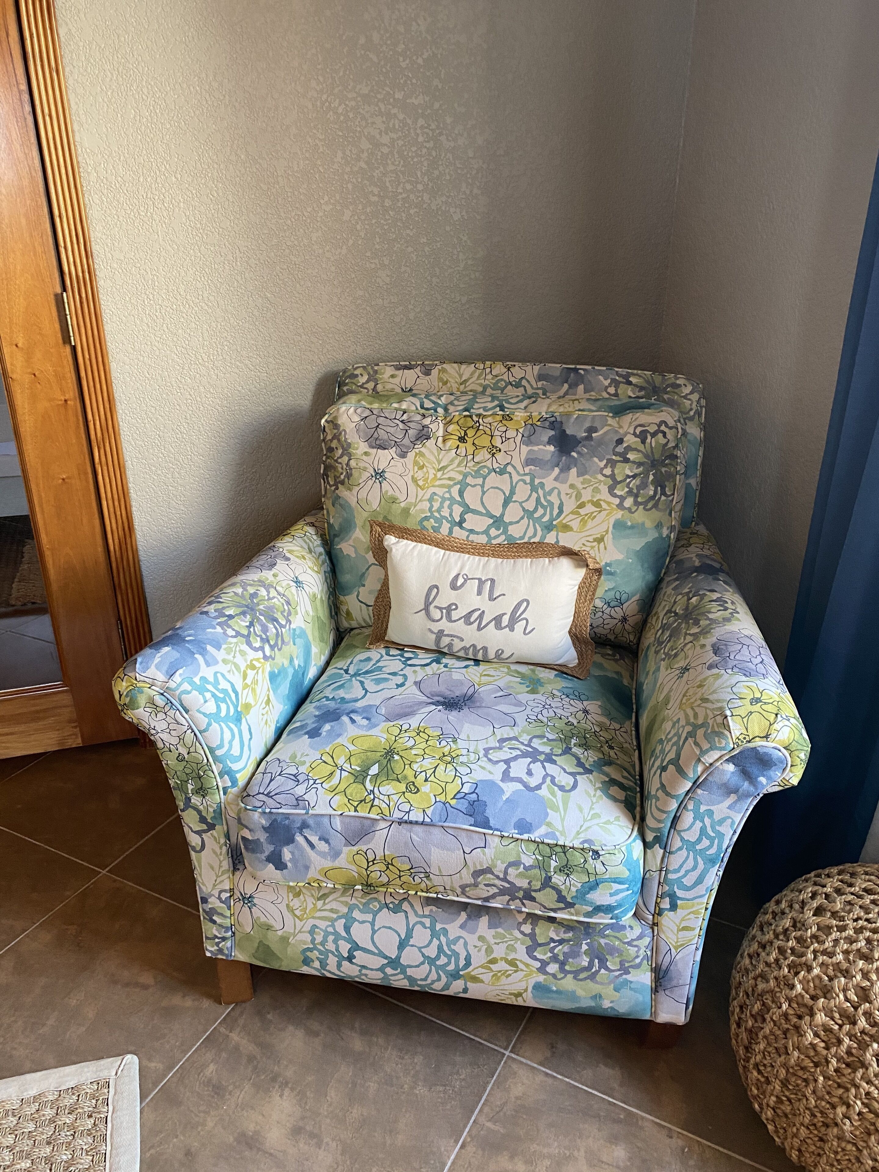 A floral-patterned armchair with a cushion that reads "beach time" against a plain wall.