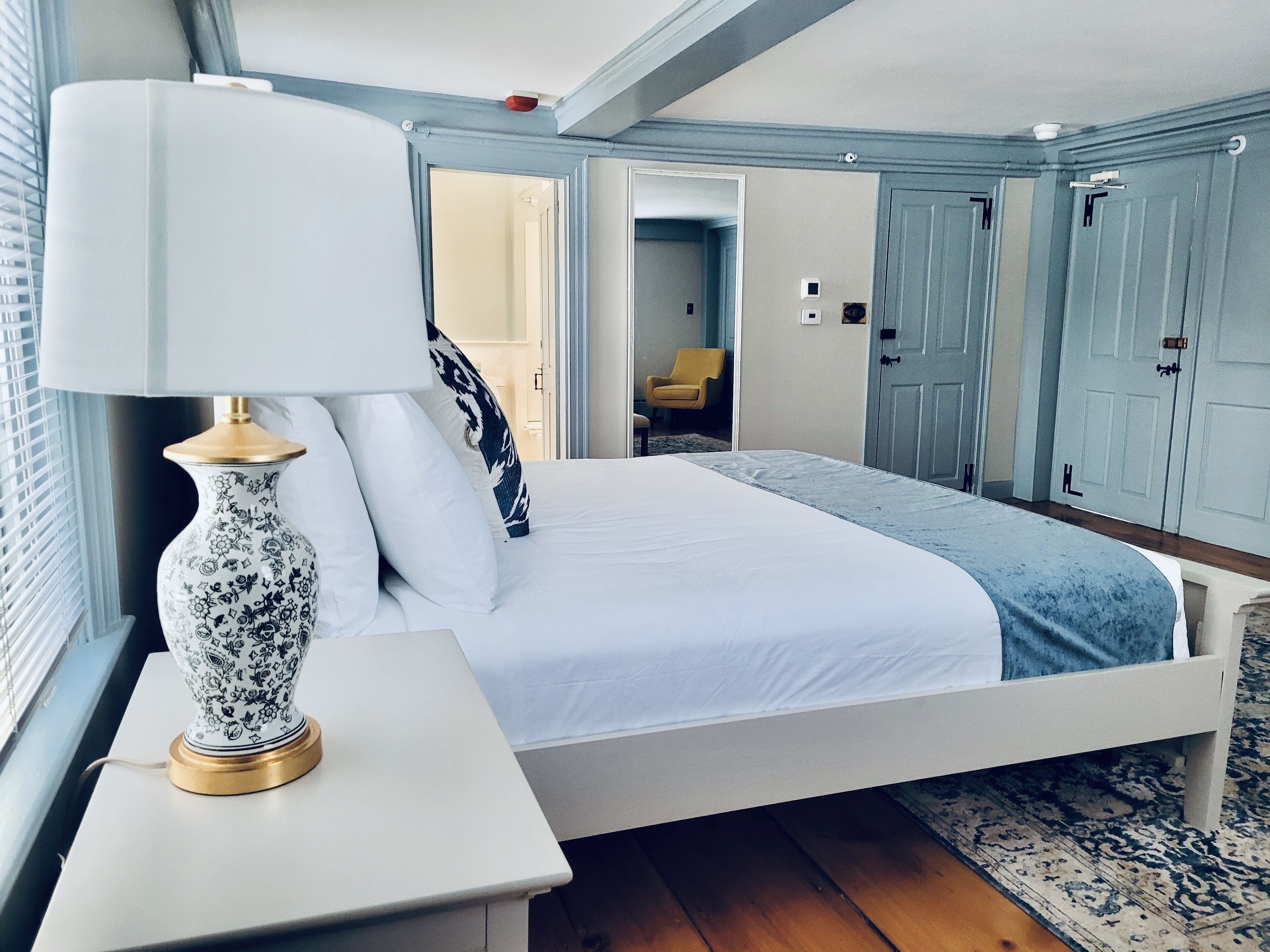 Un dormitorio con cama, una mesita de noche con lámpara, un florero estampado y una ventana con persianas.