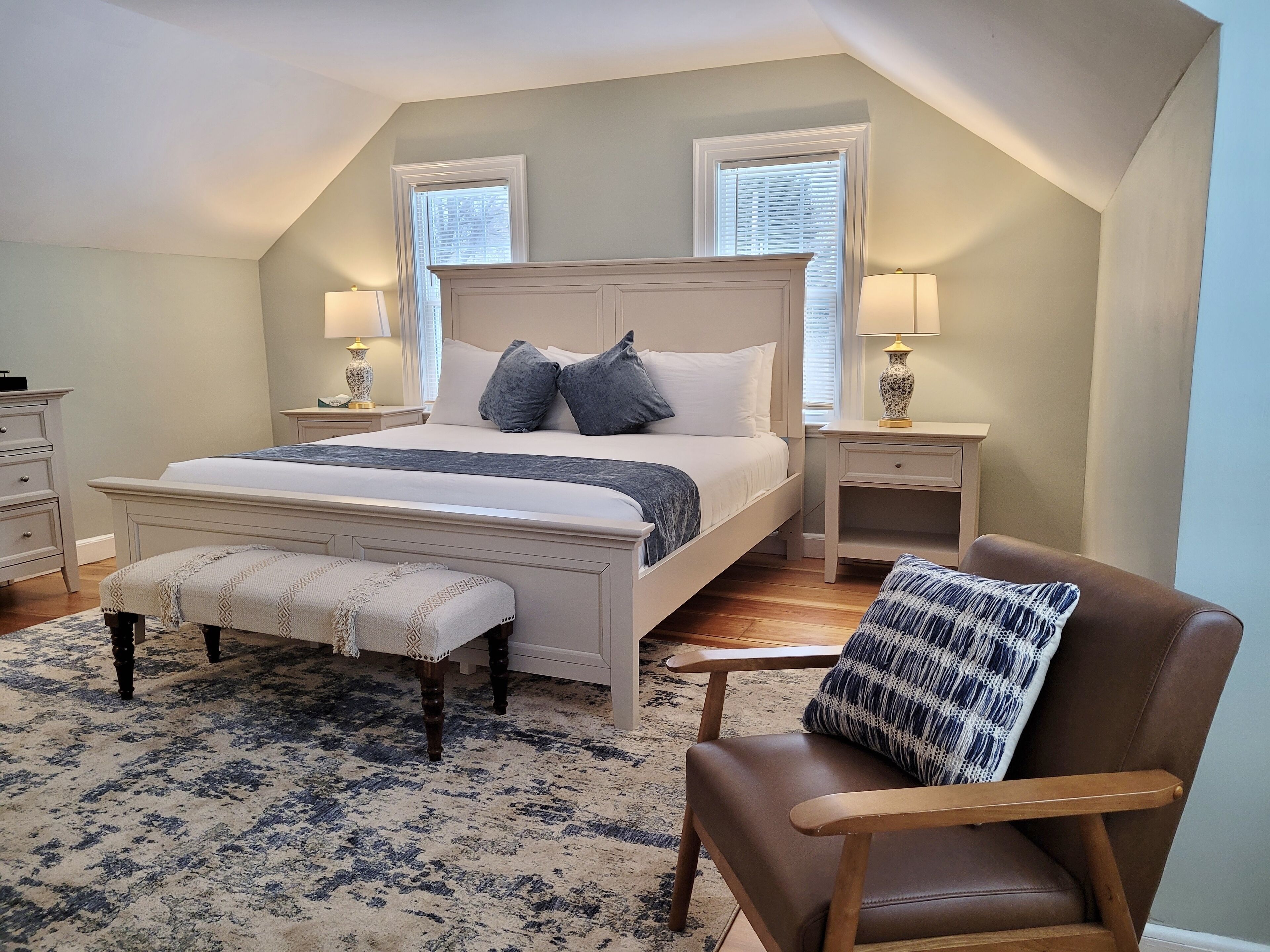 A bedroom with a large bed, two bedside tables, a bench, a chair, and a patterned rug.