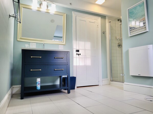 Un baño con un tocador azul oscuro, una puerta blanca, cabina de ducha con mampara de vidrio y un cuadro en la pared.