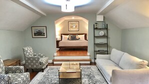 Een moderne woonkamer met een bank, fauteuil, salontafel en uitzicht op een slaapkamer met een bed en trap.