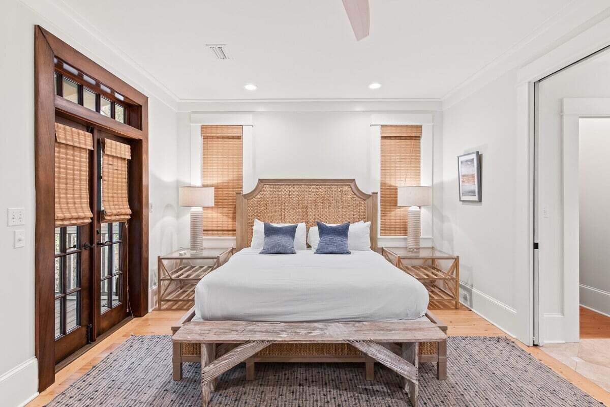 A bedroom with a large bed, wooden headboard, two bedside tables with lamps, a bench, and a view of a balcony with wooden blinds.