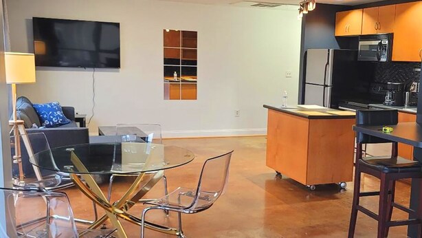 A modern kitchen with a central island, a dining area with a glass table and metal chairs, a flat-screen TV mounted on the wall, and a ceiling fan.