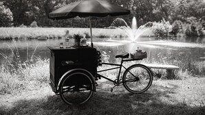 Un vélo stationnaire monté sur un chariot et doté d’un auvent, placé près d’un étang avec une fontaine, entouré d’herbe et d’arbres.