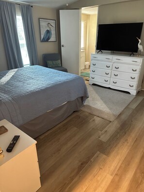 Et soveværelse med seng, kommode, fjernsyn og en dør til et andet rum.