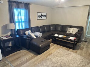 Un salon moderne avec un canapé d’angle, une table basse, une lampe et un panneau d’accueil accroché au mur.