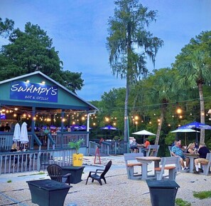 Ein Außenbereich mit Tischen, Stühlen und einer Bar. An der Bar ist ein Schild mit der Aufschrift 'SWAMPY'S BAR & GRILLE' zu sehen. Über dem Bereich hängen Lichterketten.