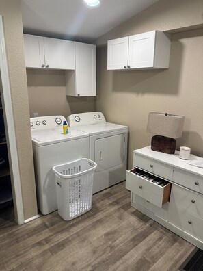 Une buanderie équipée d’un lave-linge et d’un sèche-linge, d’un panier à linge blanc et d’un meuble sur lequel est posée une lampe.