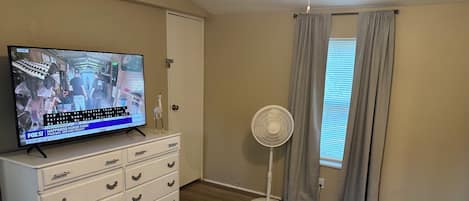 Une chambre à coucher comprenant un lit, une commode, un téléviseur diffusant un programme d’information, un ventilateur sur pied et une fenêtre avec des rideaux.