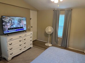 Une chambre à coucher comprenant un lit, une commode, un téléviseur diffusant un programme d’information, un ventilateur sur pied et une fenêtre avec des rideaux.