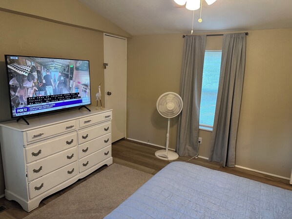 A bedroom with a bed, a dresser, a television displaying a news program, a standing fan, and a window with curtains. - “Divers Inn” on The Rainbow River (Dunnellon)