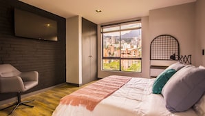 Une chambre moderne avec un grand lit, un fauteuil de bureau, un téléviseur fixé au mur et une fenêtre donnant sur la ville.