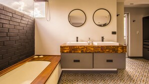 Une salle de bain moderne avec un mur en briques noires, une baignoire en bois, deux vasques avec miroirs et des carreaux de sol à motifs.
