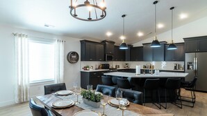 A modern kitchen with dark cabinetry, a central island, pendant lighting, and a dining area with a wooden table set for six.