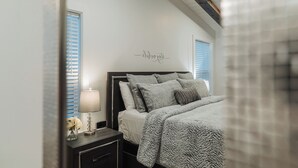 A bedroom with a sloped ceiling, a bed with grey bedding, a bedside table with a lamp, and a wall with a decorative phrase.