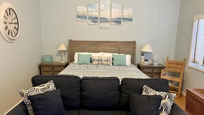 Een slaapkamer met een bed, twee nachtkastjes, een klok, een muur met kunst, een donkere bank met gestreepte kussens, een houten stoel, en een raam met zonwering.