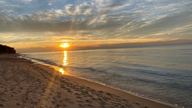 Een strand met voetafdrukken die naar het water leiden, een zonsondergang op de achtergrond en een heldere lucht.