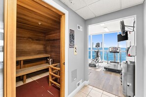 Un sauna avec des bancs en bois et un tapis rouge, un tapis de course et une vue sur la mer grâce à de grandes fenêtres.