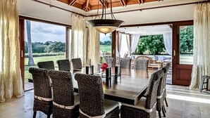 Un'ampia sala da pranzo con un tavolo grande e sedie in vimini, ampie finestre con tende e vista sull'esterno.