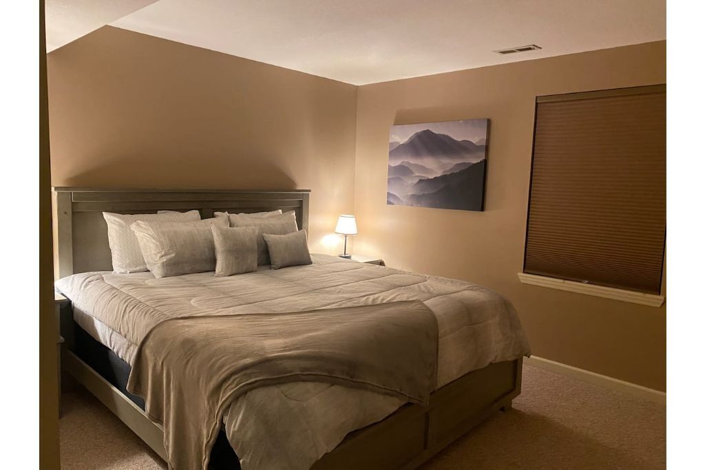 A bedroom with a bed, a nightstand, a lamp, a painting, and a window with blinds.