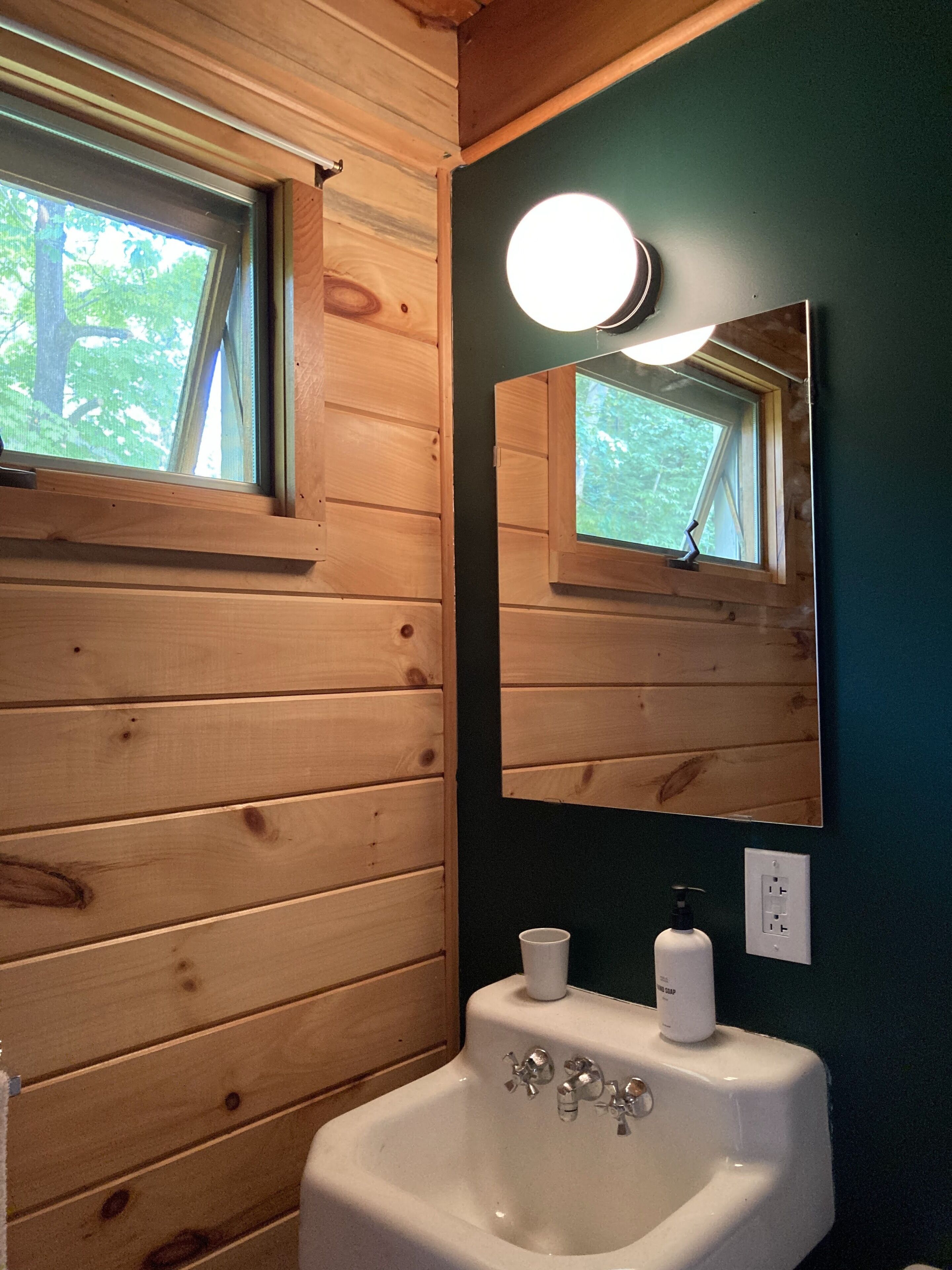 A wooden cabin bathroom with a window, a sink, a mirror, and a wall-mounted light fixture.