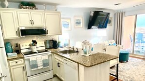 A compact kitchen with white cabinets, a microwave, stove, and a sink. There's a dining area with a chair and a small table. A TV is mounted on the wall, and there's a large window offering a view. - Amazing Unobstructed Beach Views! 🏖️Beautifully equipped with nice furnishings! (Gulf Shores)