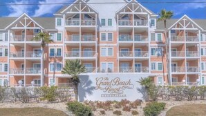 A multi-story hotel with balconies, a sign reading "The Grand Beach Resort Condominium," and a clear blue sky. - Amazing Unobstructed Beach Views! 🏖️Beautifully equipped with nice furnishings! (Gulf Shores)