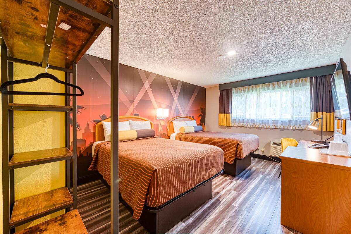 Ein Hotelzimmer mit zwei Betten, einem Schreibtisch, einem Stuhl, einem Kleiderhaken und einem Fenster mit Vorhängen.