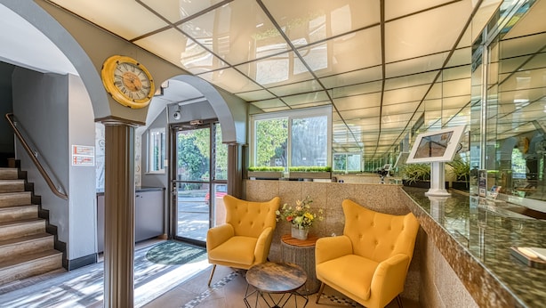 A reception area with a glass ceiling, a clock, two yellow armchairs, a small table, and a reception desk. - La Mirage Inn - Hollywood (Los Angeles)