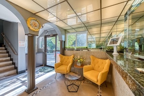 A reception area with a glass ceiling, a clock, two yellow armchairs, a small table, and a reception desk. - La Mirage Inn - Hollywood (Los Angeles)