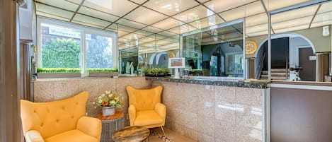 A lobby with a reception desk, two yellow armchairs, a small round table with a vase of flowers, and a tiled floor with a patterned border.