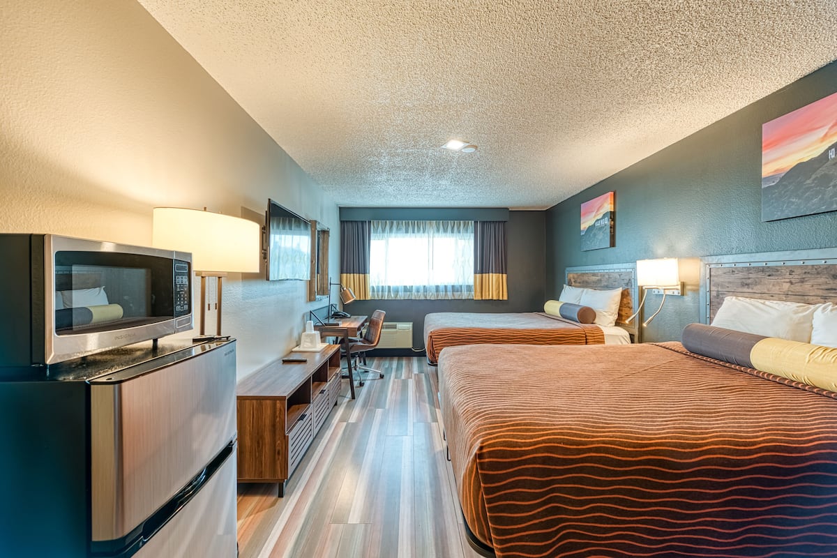 Ein Hotelzimmer mit Küchenzeile, zwei Betten, einem Schreibtisch mit Stuhl, einem Fernseher und einem großen Fenster mit Vorhängen.