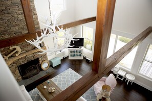 A high-ceilinged room with a stone fireplace, a white chandelier, and a cozy seating area with a small table and chairs.