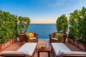 Een terras met rieten meubels, een tafel met fruit en een glas, en uitzicht op zee.