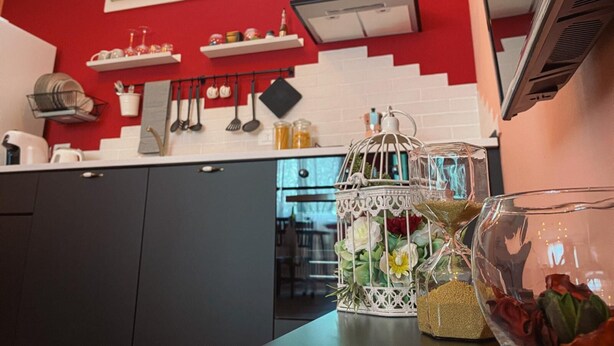 A modern kitchen with red walls, white cabinets, and a black range hood. There are hanging utensils, a white birdcage with flowers, and a glass jar with grains. - WellHome   "Barbera" Apartment (ASTI)