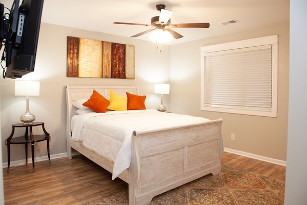 Quarto com cama, duas luminárias de cabeceira, ventilador de teto, um quadro e uma janela com persianas.