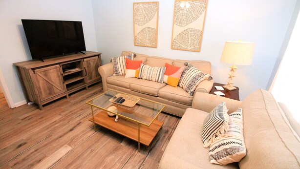A living room with a flat-screen TV, wooden entertainment unit, beige sofa with colorful cushions, a glass coffee table, and wall art. - Charming and Newly Remodeled Vacation Townhouse Located In The Secluded Sea Cabins Of Miramar Beach (Miramar Beach)