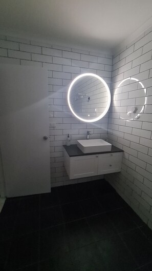A bathroom with white subway tiles, a white sink with a black countertop, and two round mirrors with illuminated frames. - Emthree Seaside Apartments (North Ward)