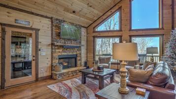 A cozy cabin interior with a stone fireplace, wooden walls, a large window, and a comfortable seating area.
