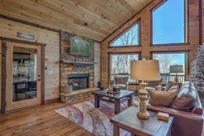 A cozy cabin interior with a stone fireplace, wooden walls, a large window, and a comfortable seating area. - Blue Ridge Cabin, Mountain Views, Hot Tub, Game Room – Sleeps 12 Near Downtown (Morganton)