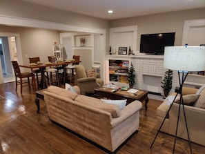 A living room with a wooden floor, beige sofa, dining table, and a TV mounted on the wall.